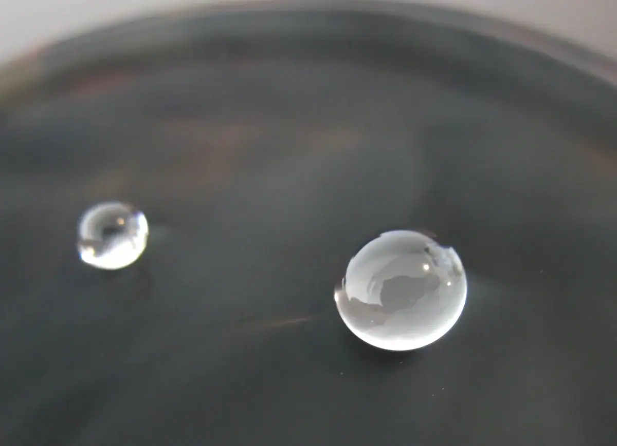Goutte d'eau suspendue sur une surface sombre, révélant des détails microscopiques et la réfraction de la lumière.