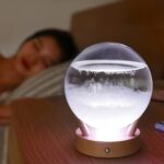 Boule de cristal illuminée avec lumière LED, posée sur une table en bois, avec une femme endormie en arrière-plan dans une chambre.