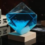 Panneau de verre en forme de cube contenant un liquide bleu avec une poudre blanche au fond, posé sur un socle en bois, illustrant un objet scientifique décoratif.