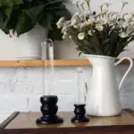 Deux vases de laboratoire en verre avec bases en bois noir, utilisés comme objets décoratifs, placés sur une table en bois avec un bouquet de fleurs blanches dans une cruche en céramique blanche.