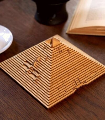 Modèle de pyramide en bois gravée avec des motifs scientifiques, posé sur une table en bois avec un livre ouvert et une tasse à café à proximité.