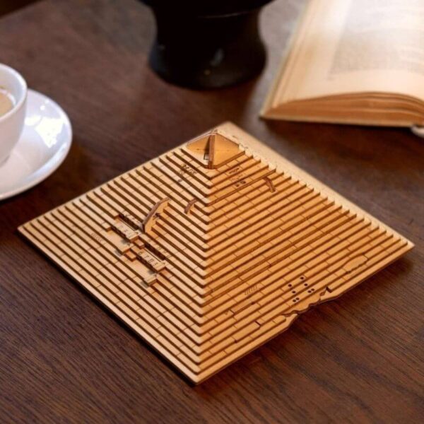 Modèle de pyramide en bois gravée avec des motifs scientifiques, posé sur une table en bois avec un livre ouvert et une tasse à café à proximité.