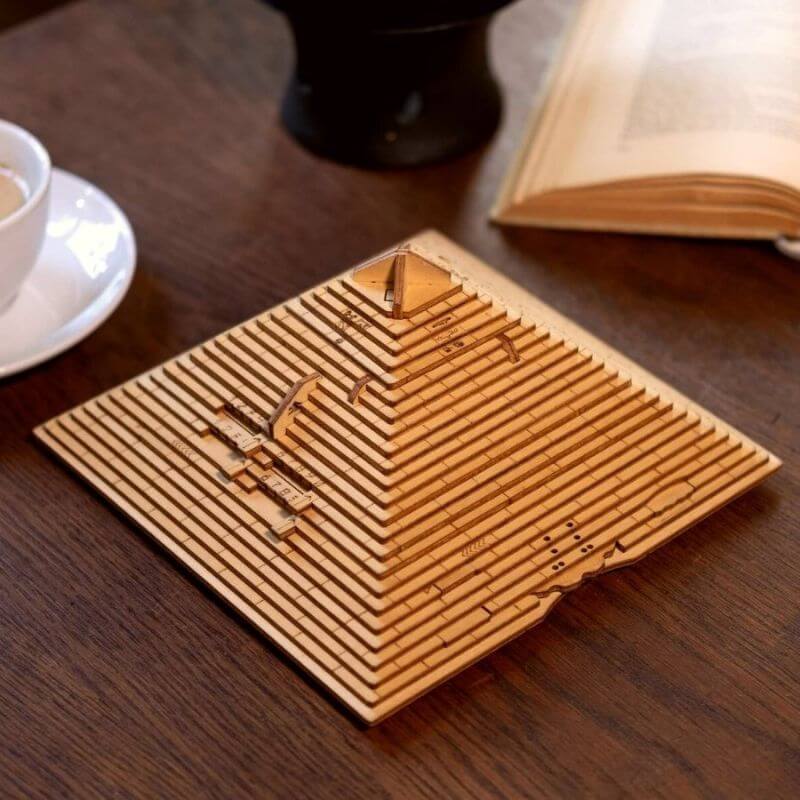 Modèle de pyramide en bois gravée avec des motifs scientifiques, posé sur une table en bois avec un livre ouvert et une tasse à café à proximité.
