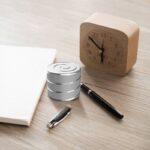 Horloge en bois avec aiguilles noires, bloc-notes blanc, stylo et boîte métallique sur une surface en bois clair.