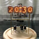 Horloge à affichage numérique vintage avec tubes en verre orange affichant l'heure 20:13:00, sur un socle en bois, dans un décor industriel.