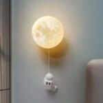 Lampe murale en forme de lune avec figurine d'astronaute suspendue, créant une ambiance spatiale et chaleureuse dans la pièce.