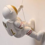 Figurine d'astronaute en plastique blanc avec détails dorés et rouges, suspendue à une corde, évoquant l'exploration spatiale.