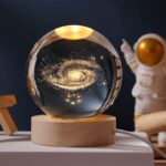 Boule en verre contenant une galaxie miniature avec étoiles et poussière cosmique, posée sur un socle en bois, idéale pour la décoration d'intérieur à thème scientifique ou cosmologique.