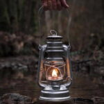 Lampe à pétrole en métal avec flamme allumée, suspendue au-dessus d'une rivière dans un environnement forestier.