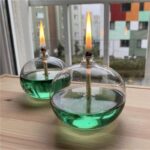 Bocal en verre contenant un liquide vert avec une mèche enflammée, placé sur une surface en bois près d'une fenêtre.
