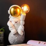 Figurine d'astronaute en costume avec une lampe LED allumée, posée sur une surface avec un décor de style lunaire. Idéal pour décoration scientifique ou thématique spatiale.