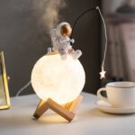 Lampe de nuit en forme de lune avec figurine d'astronaute assis dessus, éclairage doux et ambiance spatiale, décoration originale pour chambre d'enfant ou espace de rêve.