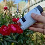 Petite lunette de microscope portable utilisée pour observer de près des fleurs rouges dans un jardin urbain, avec un fond flou de bâtiments modernes et de végétation.