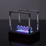 Pendule de Newton illuminée avec sphères suspendues et base lumineuse colorée, exposée dans un environnement sombre pour mettre en valeur l'effet de mouvement et de lumière.
