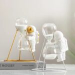 Horloge décorative avec deux figurines astronautes en plastique blanc, tenant des ballons en verre, sur une étagère avec un livre de science.