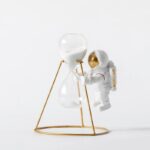 Vase décoratif en forme de sablier avec un astronaute miniature, contenant du sable blanc, sur un support en métal doré, idéal pour la décoration scientifique.