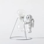 Sablier en verre avec support métallique, décor de style scientifique et spatial, représentant un astronaute en combinaison blanche manipulant un sablier.