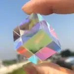 Cristal en forme de cube multifacette, transparent avec reflets colorés, tenu entre deux doigts contre un ciel bleu.