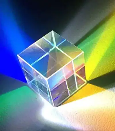 Cube en verre transparent projetant des lumières colorées bleues, vertes, jaunes et roses, créant un jeu de reflets et d'ombres sur une surface sombre.