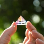 Pyramide en cristal transparent tenue entre deux doigts, reflet de lumière colorée, fond flou de nature verte.