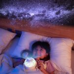 Enfant souriant dans une chambre, utilisant une lampe projection d'étoiles pour découvrir le ciel nocturne depuis son lit. Idéal pour éveiller la curiosité scientifique.