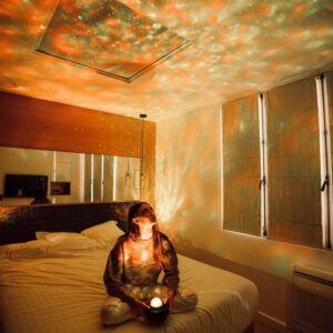 Femme assise sur un lit dans une chambre, utilisant une boule à étoiles pour projeter des motifs lumineux sur les murs et le plafond, créant une ambiance relaxante et magique.