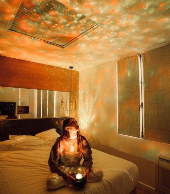 Femme assise sur un lit dans une chambre, utilisant une boule à étoiles pour projeter des motifs lumineux sur les murs et le plafond, créant une ambiance relaxante et magique.