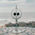 Globe en verre avec deux aimants noirs suspendus à l'intérieur, posé sur une surface en mosaïque, avec vue sur la mer et des maisons en arrière-plan.
