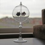Vase en verre transparent contenant deux petits objets noirs en forme de carrés, placé sur une table près d'une fenêtre avec un fond flou urbain.