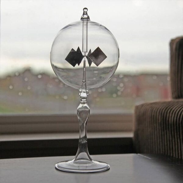 Vase en verre transparent contenant deux petits objets noirs en forme de carrés, placé sur une table près d'une fenêtre avec un fond flou urbain.