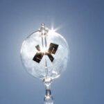 Horloge solaire en verre transparent avec cadran solaire intégré, suspendue dans le ciel en plein jour, reflétant la lumière du soleil.