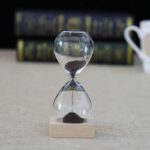 Sablier de laboratoire en verre avec sable noir, symbole de la mesure du temps dans la science, posé sur une base en bois, avec un fond flou de livres scientifiques.