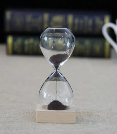 Sablier de laboratoire en verre avec sable noir, symbole de la mesure du temps dans la science, posé sur une base en bois, avec un fond flou de livres scientifiques.