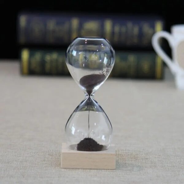 Sablier de laboratoire en verre avec sable noir, symbole de la mesure du temps dans la science, posé sur une base en bois, avec un fond flou de livres scientifiques.