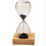Sablier en verre avec sable noir, symbole du temps qui passe, posé sur une base en bois clair, idéal pour la décoration scientifique ou éducative.