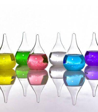 Vases de laboratoire en verre avec liquides colorés jaune, vert, rose, transparent, bleu et violet, disposés en ligne avec leur reflet sur une surface lisse.