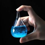Vial de laboratoire contenant un liquide bleu lumineux, tenu entre le pouce et l'index d'une main humaine, contre un fond noir.