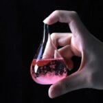Pipette de laboratoire contenant un liquide rose effervescent, tenue par une main humaine, sur fond noir. Idéal pour la décoration scientifique ou éducative.