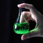 Petite fiole de laboratoire contenant un liquide vert fluorescent, tenue par une main, sur fond noir.