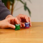 Puce de verre colorée tenue entre les doigts, posée sur une surface en bois, avec un fond flou de plantes vertes.