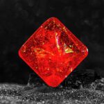 Pierre précieuse rouge en forme de cube, éclatante et brillante, avec des reflets dorés et une surface polie, posée sur une surface sombre pour mettre en valeur sa couleur vive.