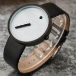 Montre minimaliste avec cadran blanc, aiguilles noires et petite touche noire, bracelet en silicone noir, design épuré et moderne.