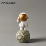 Figurine d'astronaute en plastique blanc avec casque doré brillant, posée sur une petite lune en pierre.