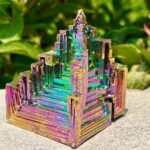 Pièce décorative en métal iridescent, inspirée d'une structure géométrique pyramidale, avec des reflets colorés, posée sur une surface rocheuse en extérieur.