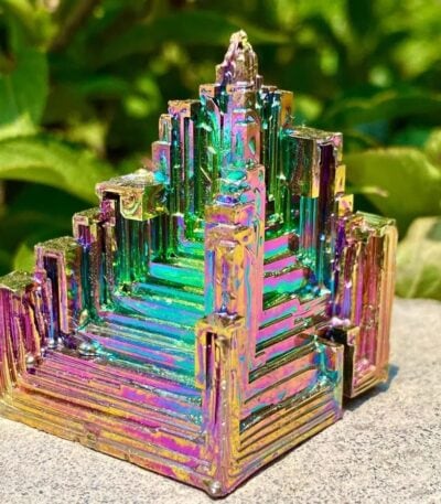 Pièce décorative en métal iridescent, inspirée d'une structure géométrique pyramidale, avec des reflets colorés, posée sur une surface rocheuse en extérieur.