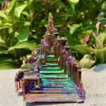 Pyramide en cristal irisé avec reflets multicolores, posée sur une surface en pierre, entourée de végétation verte en arrière-plan.