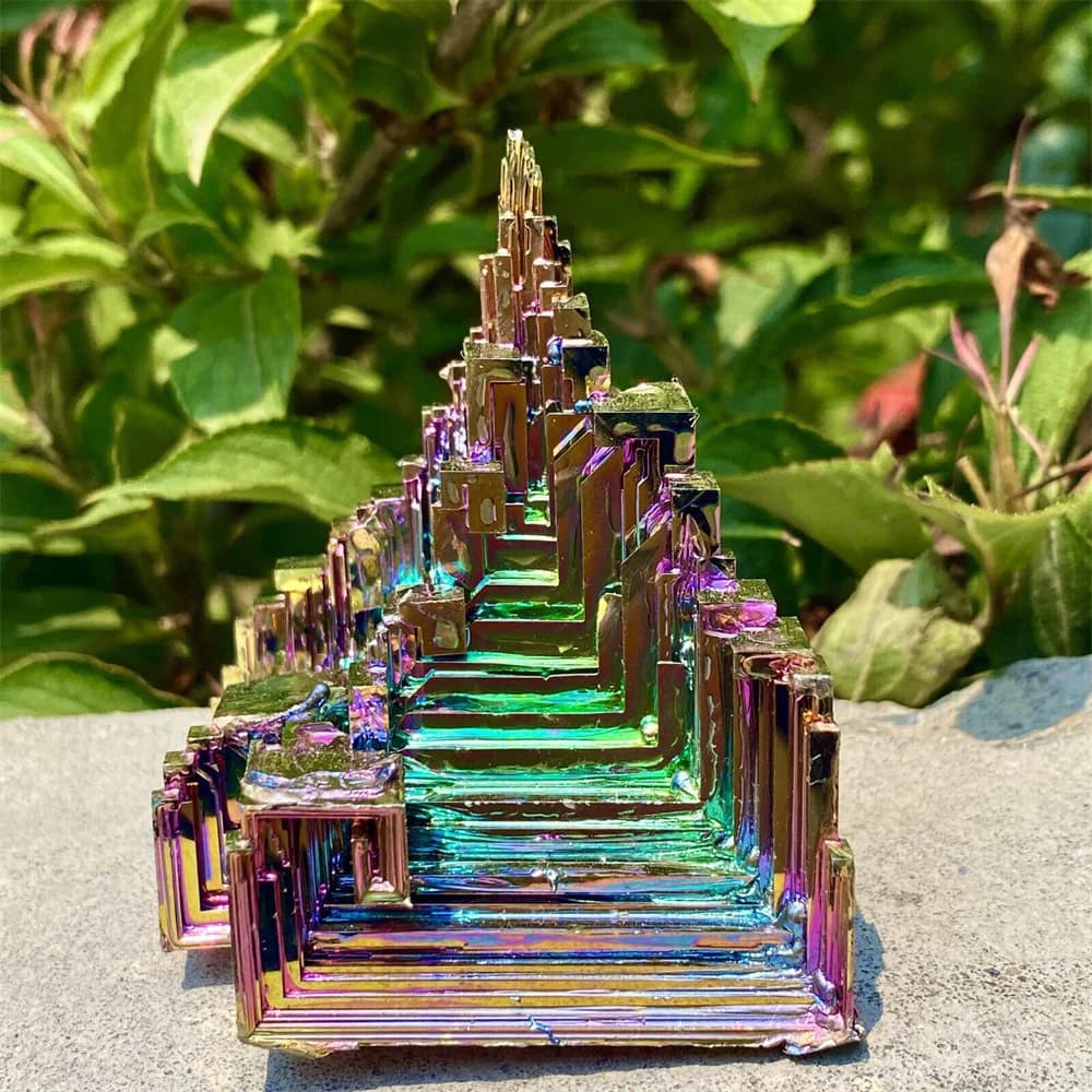 Pyramide en cristal irisé avec reflets multicolores, posée sur une surface en pierre, entourée de végétation verte en arrière-plan.