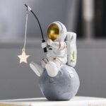 Figurine d'astronaute en plastique blanc avec casque doré, assis sur une petite lune grise, tenant une canne à pêche avec une étoile suspendue, décor de style scientifique et spatial.