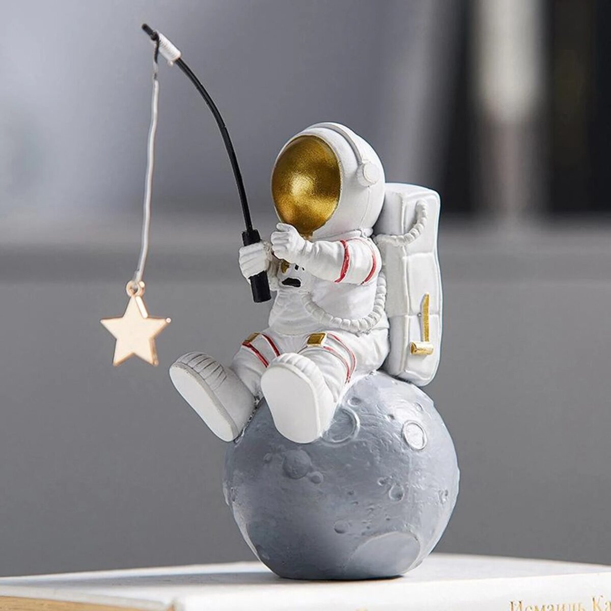 Figurine d'astronaute en plastique blanc avec casque doré, assis sur une petite lune grise, tenant une canne à pêche avec une étoile suspendue, décor de style scientifique et spatial.