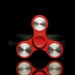 Fidget spinner rouge avec trois bras en métal brillant, conçu pour la détente et le divertissement, posé sur un fond noir pour mettre en valeur ses détails.
